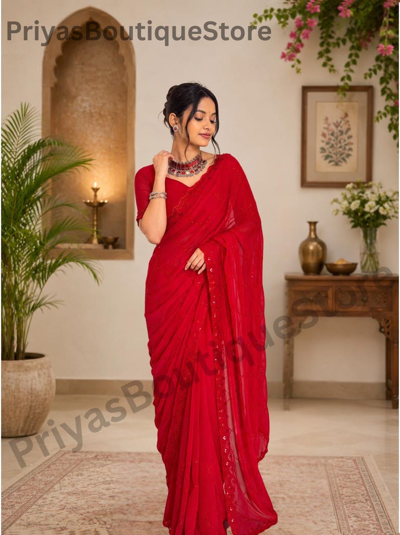 May include: A vibrant red saree featuring a lightweight, flowing fabric with subtle sequin embellishments along the border. The garment is paired with a matching short-sleeved blouse and a metallic choker necklace, creating a cohesive ensemble.