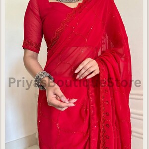 May include: A vibrant red saree featuring sheer fabric with delicate sequin embroidery in a geometric pattern. The garment includes a matching short-sleeved blouse with embellished edges, paired with a silver-toned choker and a wide cuff bracelet.