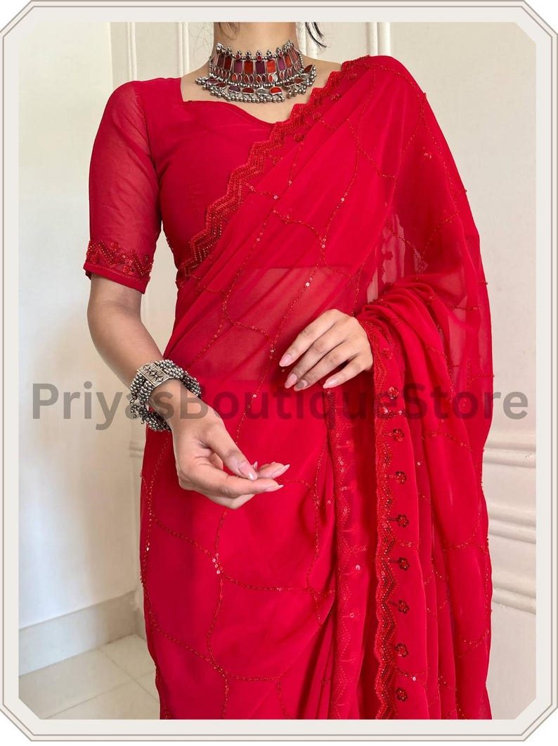 May include: A vibrant red saree featuring sheer fabric with delicate sequin embroidery in a geometric pattern. The garment includes a matching short-sleeved blouse with embellished edges, paired with a silver-toned choker and a wide cuff bracelet.