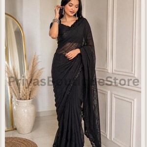 May include: A black saree featuring a sheer fabric with intricate tonal embroidery and sequin embellishments along the border and pallu. The garment is paired with a matching black short-sleeved blouse. The text PriyasBoutiqueStore is visible.