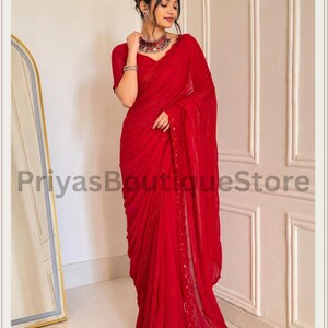 May include: A vibrant red saree featuring delicate embroidery along the border and subtle sequin embellishments throughout the fabric. The garment is paired with a matching short-sleeved blouse, creating a cohesive and elegant ensemble.