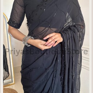 May include: A charcoal black saree featuring a sheer, lightweight fabric with an intricate, repeating lattice pattern created by small black beads and sequins. The garment includes a matching blouse with short sleeves and a scalloped, beaded neckline.