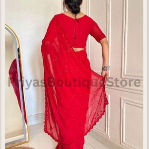 May include: A vibrant red saree featuring a sheer fabric with delicate sequin embroidery in a wavy pattern and a scalloped border. The ensemble includes a matching red blouse with short sleeves and a button-down back detail. Text reads PriyasBoutiqueStore.