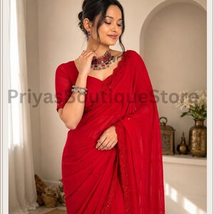 May include: A vibrant red saree featuring delicate embroidery along the border, paired with a matching short-sleeved blouse. The ensemble is accessorized with a silver-toned necklace and stacked bracelets, creating a cohesive and elegant appearance.