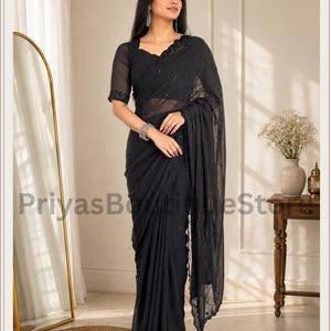 May include: A black saree featuring a sheer fabric with intricate tonal embroidery and subtle sequin embellishments. The garment includes a matching short-sleeved blouse with a scalloped neckline and a draped pallu that falls gracefully to the floor.