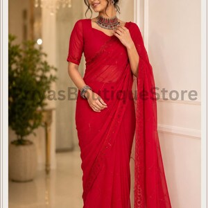 May include: A vibrant red saree featuring a sheer, lightweight fabric with delicate tonal embroidery and subtle sequin embellishments along the border. The garment includes a matching short-sleeved blouse and a long, flowing drape.