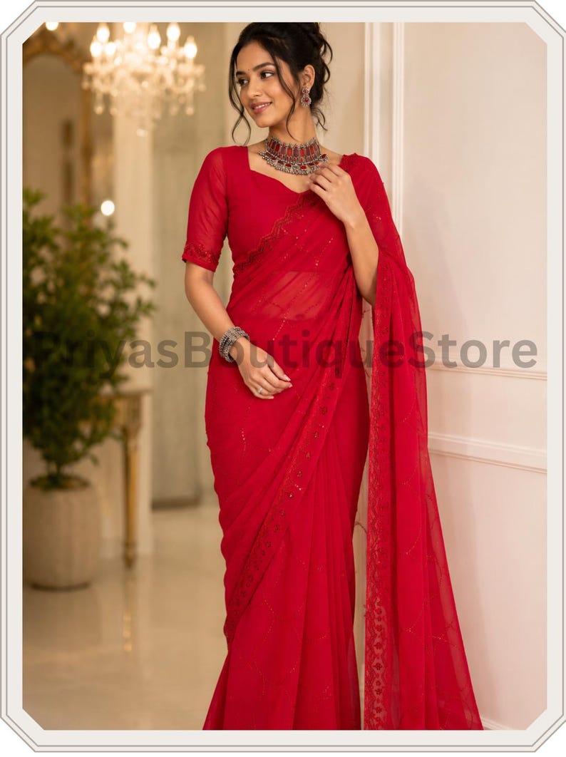 May include: A vibrant red saree featuring a sheer, lightweight fabric with delicate tonal embroidery and subtle sequin embellishments along the border. The garment includes a matching short-sleeved blouse and a long, flowing drape.