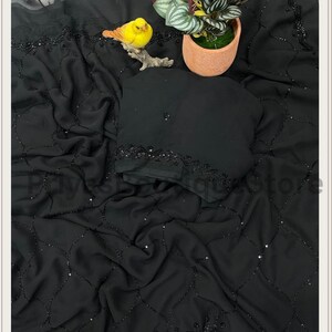 May include: A black chiffon saree featuring a delicate lattice pattern created with small black beads and sequins. The fabric is draped in soft folds, showcasing a decorative border with intricate beadwork and a matching black blouse piece.