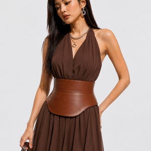 Puede incluir: Un vestido marrón oscuro con cuello halter, escote en V profundo y falda plisada fluida. El vestido se ajusta a la cintura con un cinturón ancho y estructurado tipo corsé de piel sintética marrón. La tela parece ligera y semitransparente.