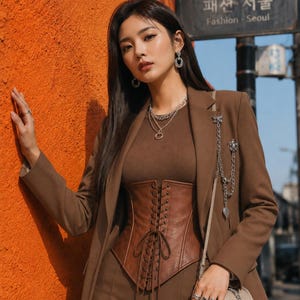 May include: A brown blazer worn over a brown ribbed top and a brown lace-up corset. The outfit includes a silver chain accessory pinned to the lapel, a layered silver necklace, silver earrings, multiple silver rings, and a beige quilted handbag.