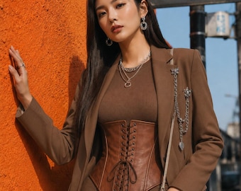 May include: A brown blazer worn over a brown ribbed top and a brown lace-up corset. The outfit includes a silver chain accessory pinned to the lapel, a layered silver necklace, silver earrings, multiple silver rings, and a beige quilted handbag.