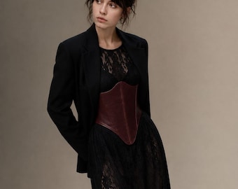 Può includere: Un blazer nero indossato sopra un abito in pizzo nero con un corsetto bordeaux scuro. L'abito presenta una gonna lunga con un alto spacco laterale, mentre il blazer strutturato ha maniche lunghe e un classico colletto a revers.