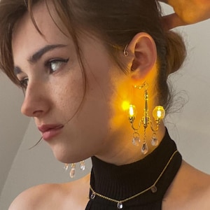 Puede incluir: Una persona lleva un top negro de tirantes con un collar de cadena dorada y pendientes de araña con gotas de cristal transparente. Los pendientes están iluminados con pequeñas luces amarillas.