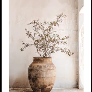 May include: A large, weathered, tan-colored earthenware vessel with a wide, rounded body and a narrow neck. The vessel features a textured, mottled surface with dark brown patches. A thin, bare-branched plant with small, dried leaves sits inside.