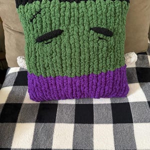 Puede incluir: Un cojín cuadrado de ganchillo con un rostro verde, cabello negro, cejas negras y una pequeña cicatriz negra cosida en la frente. El cojín tiene una base morada, dos pequeños pernos laterales de color gris claro y una textura de hilo grueso.