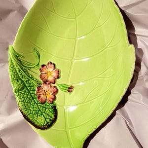 May include: A glossy light green ceramic dish shaped like a large leaf with embossed veins. A textured green leaf section is attached to the left side, featuring two small pink flowers with yellow centres and brown details on the petals.