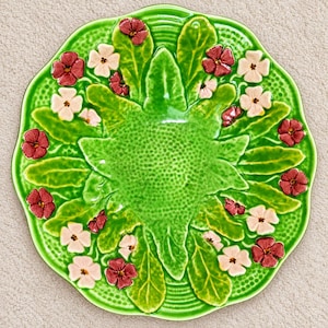 Op de afbeelding: Een ronde keramische plaat met een geschulpte rand, voorzien van een levendig groen geglazuurd oppervlak versierd met reliëf groene bladeren en een verspreiding van kleine, verhoogde vijfbladige bloemen in donkerrood en lichtroze.