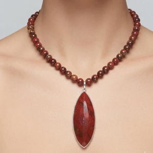 Op de afbeelding: Een ketting met een streng ronde, roodbruine kralen en een grote, gefacetteerde, marquisevormige hanger van een dieprode, gevlekte steen. De hanger is gezet in een eenvoudige, zilverkleurige metalen rand, wat zorgt voor een robuuste uitstraling.