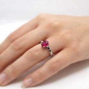 Op de afbeelding: Een zilveren ring met een centrale ovaal geslepen, levendig rode edelsteen die door vier pootjes wordt vastgehouden. De band is versierd met kleine, donkere, marquisevormige steentjes aan de zijkanten voor een verfijnde uitstraling.
