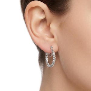 May include: A silver-toned hoop earring featuring a textured, braided metal design. The earring is circular in shape with a hinged clasp mechanism at the top, securing the hoop through the earlobe. The metal surface has a polished, reflective finish.