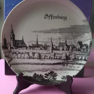 May include: A white ceramic decorative plate featuring a black ink illustration of the city of Offenburg. The scene depicts a historic town skyline with towers and buildings behind a wall, with the text Offenburg and Kinzig flu printed.