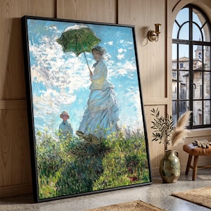 May include: A framed impressionist painting depicting a person in a white dress holding a green parasol, standing in a grassy field with a child nearby under a bright blue sky with white clouds. The artwork is displayed in a black frame.
