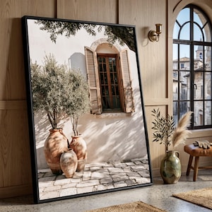 May include: A framed wall art print depicting a Mediterranean scene with three terracotta pots containing olive trees placed against a white wall with a wooden shuttered window. The scene features a stone-paved ground and tree branch shadows.
