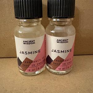 May include: Two small clear glass bottles of Jasmine fragrance oil with black ribbed screw caps. Each white label features the text Ancient Wisdom, Jasmine, Fragrance Oil, Made in England, and 10 ml / 0.33 fl oz, with pink and brown geometric accents.