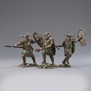 Puede incluir: Tres figuras de guerreros en miniatura con cascos con cuernos, armadura de cuero marrón y túnicas verdes. Cada figura se apoya sobre una base rocosa, portando una lanza, una maza o un escudo con un diseño de nudos circulares.