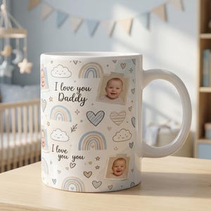 May include: White ceramic mug with the text "I love you Daddy" and baby photos, decorated with pastel rainbows, clouds, and hearts. The mug is on a wooden table in a nursery setting.