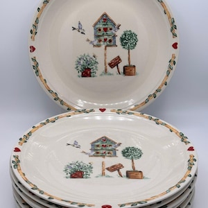 May include: A stack of cream-colored ceramic plates featuring a central illustration of a blue birdhouse, a potted topiary, and a small sign. The rim is decorated with a brown vine pattern interspersed with small red hearts around the edge.