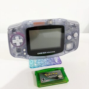 Puede incluir: Una consola portátil Game Boy Advance de color morado translúcido con un marco de pantalla negro y botones blancos, situada sobre un cartucho de juego verde de Pokemon Emerald Version con el título y el símbolo de clasificación E.
