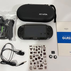 May include: A black Sony PlayStation Vita handheld console with a matching black carrying case, an AC power adapter, wired earphones, a sheet of small decorative stickers, a memory card, two paw-print thumb grips, and a tempered glass screen protector.