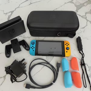 May include: A Nintendo Switch console with a blue and yellow controller, a black dock, a carrying case, a charging grip, a power adapter, an HDMI cable, and two pairs of blue and orange silicone controller covers with paw print thumb grips.
