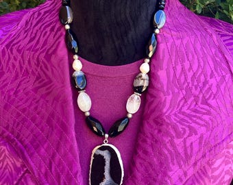 May include: A black and white agate pendant necklace with a silver-colored trim. The necklace features black and white beads, and is displayed on a black mannequin head. The mannequin is set against a purple jacket.