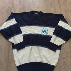 May include: A long-sleeve crewneck jumper featuring a colour-blocked design in navy blue and cream. The knit pattern includes textured sections and a circular emblem on the chest with the text "Fiers d'&ecirc;tre Marseillais" and a large letter M.