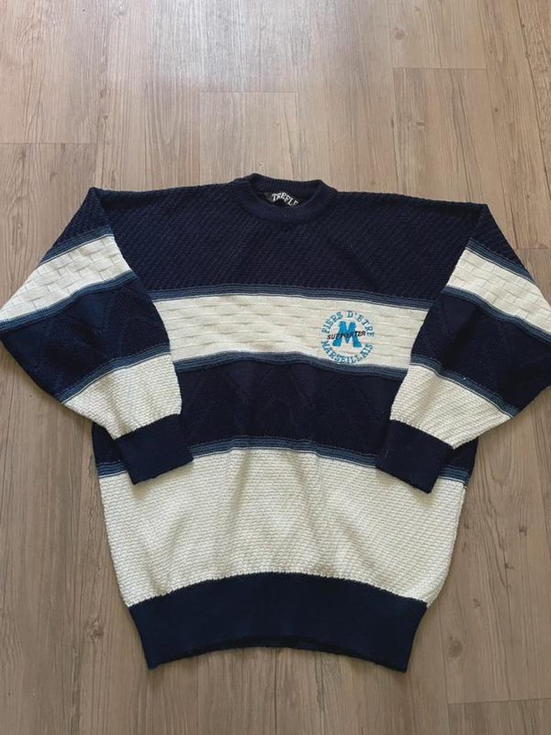 May include: A long-sleeve crewneck jumper featuring a colour-blocked design in navy blue and cream. The knit pattern includes textured sections and a circular emblem on the chest with the text "Fiers d'&ecirc;tre Marseillais" and a large letter M.