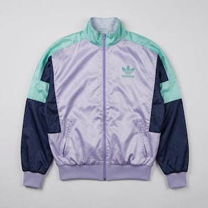 May include: A lavender, mint green, and navy blue zip-up track jacket with a stand-up collar, ribbed cuffs and hem, and two front pockets. The Adidas trefoil logo and text are embroidered in mint green on the upper left chest area.