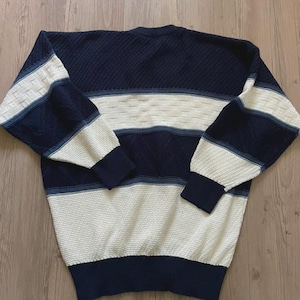 May include: A long-sleeve crew neck jumper featuring a colour-blocked design in navy blue and cream. The garment displays a textured knit pattern with horizontal stripes across the chest, sleeves, and hem, finished with solid navy ribbed cuffs.