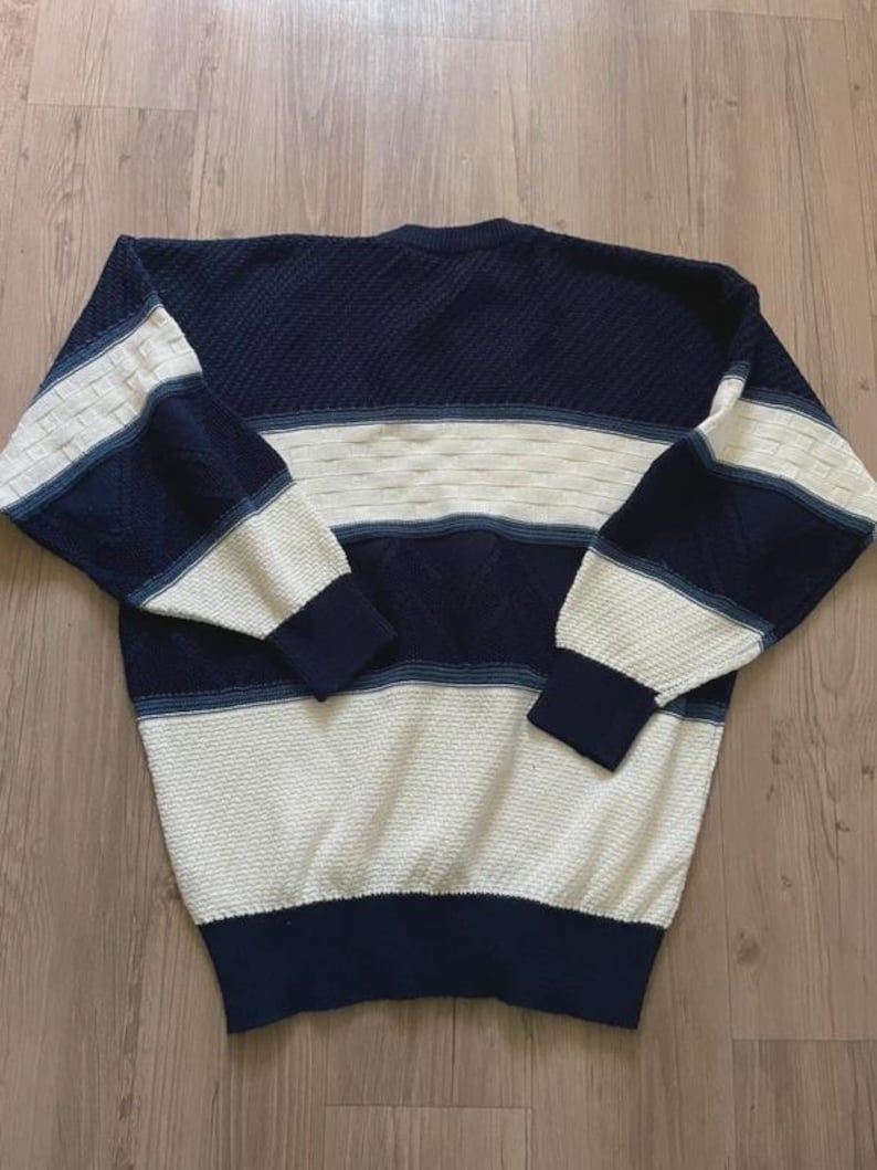 May include: A long-sleeve crew neck jumper featuring a colour-blocked design in navy blue and cream. The garment displays a textured knit pattern with horizontal stripes across the chest, sleeves, and hem, finished with solid navy ribbed cuffs.