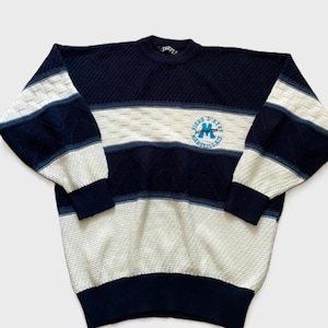 May include: A long-sleeve knit jumper featuring horizontal stripes in navy blue, white, and light blue. The jumper has a textured diamond pattern and a circular embroidered logo on the chest that reads Fiers D'Etre Marseillais with a blue letter M.