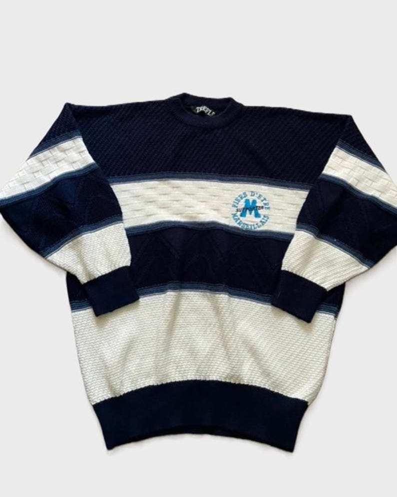 May include: A long-sleeve knit jumper featuring horizontal stripes in navy blue, white, and light blue. The jumper has a textured diamond pattern and a circular embroidered logo on the chest that reads Fiers D'Etre Marseillais with a blue letter M.