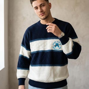 May include: A long-sleeve knit jumper featuring horizontal stripes in navy blue and white. The jumper has a textured pattern and a circular emblem on the chest with the text FIERS D'ETRE MARSEILLAIS and the word SUPPORTER in the centre.