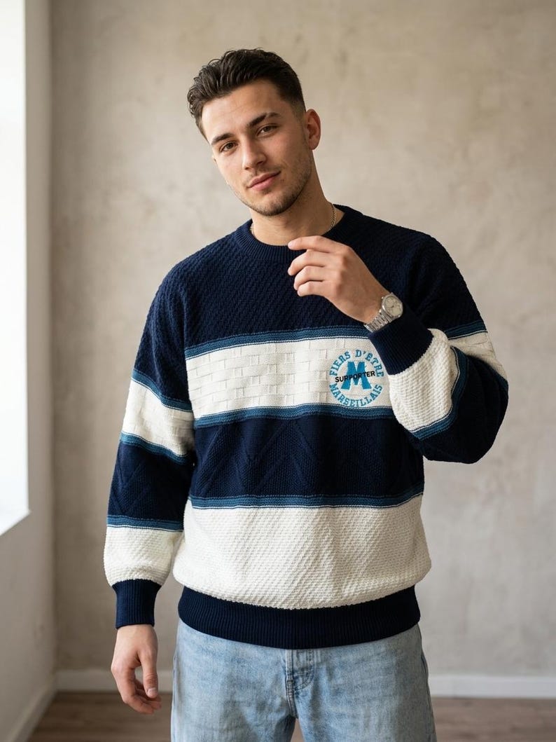 May include: A long-sleeve knit jumper featuring horizontal stripes in navy blue and white. The jumper has a textured pattern and a circular emblem on the chest with the text FIERS D'ETRE MARSEILLAIS and the word SUPPORTER in the centre.