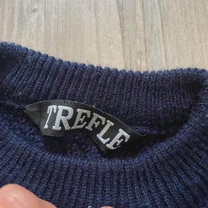 May include: A dark navy blue knit jumper featuring a ribbed crew neckline. A black fabric label is sewn into the inner collar, displaying the word TREFLE in white capital letters. The knit texture is visible throughout the garment's collar area.