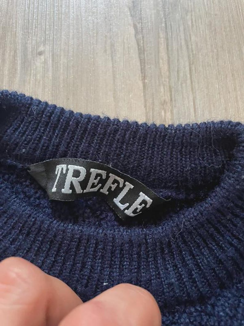 May include: A dark navy blue knit jumper featuring a ribbed crew neckline. A black fabric label is sewn into the inner collar, displaying the word TREFLE in white capital letters. The knit texture is visible throughout the garment's collar area.