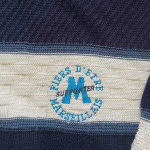 May include: A navy blue and cream knit jumper featuring a textured basketweave pattern. A circular emblem is embroidered in the centre with the light blue text FIERS D'ETRE MARSEILLAIS and the word SUPPORTER printed across a large blue letter M.