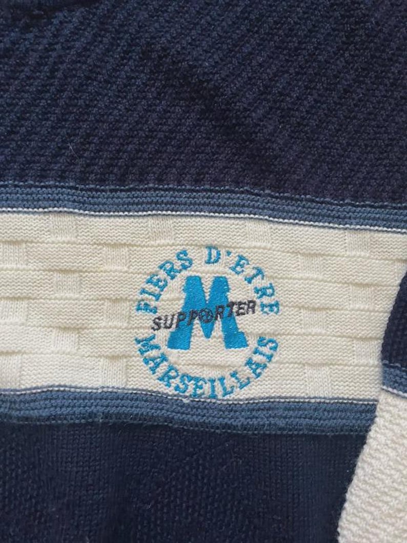 May include: A navy blue and cream knit jumper featuring a textured basketweave pattern. A circular emblem is embroidered in the centre with the light blue text FIERS D'ETRE MARSEILLAIS and the word SUPPORTER printed across a large blue letter M.