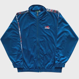 Op de afbeelding: Een blauw trainingsjack met een volledige ritssluiting en een opstaande kraag. De jas heeft wit en rood gestreepte patronen langs de schouders en mouwen. Een klein logo is zichtbaar op de borst.