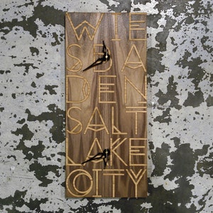 May include: A wooden clock with the words "Salt Lake City" engraved in a geometric font. The clock hands are black and the background is a light brown wood.